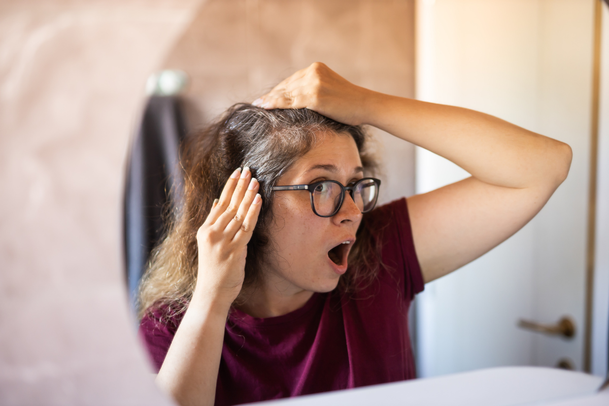 Young woman with glasses looking shocked at her premature grey hair roots in a bathroom mirror